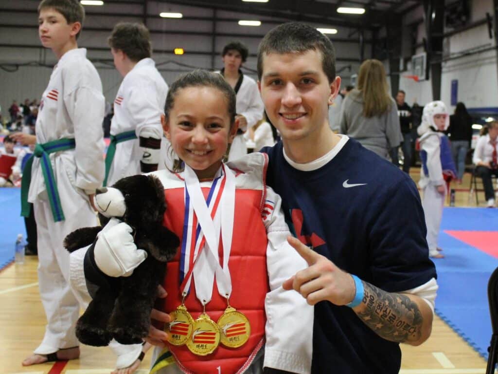 Midwest Tae Kwon Do kid in sparring gear at an event with medals and a teddy bear.